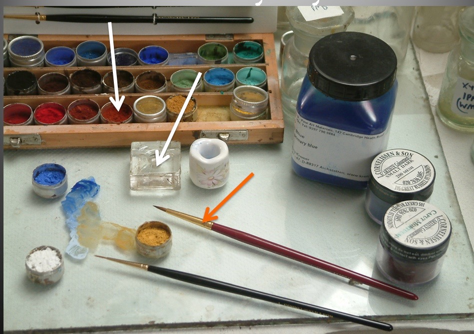 Retouching kit on a glass palette: a wooden box of dry pigments, small pots of blue, white and ochre pigment, mixed blue and yellow paint, and two fine sable brushes, with arrows highlighting the pigments and brush tip